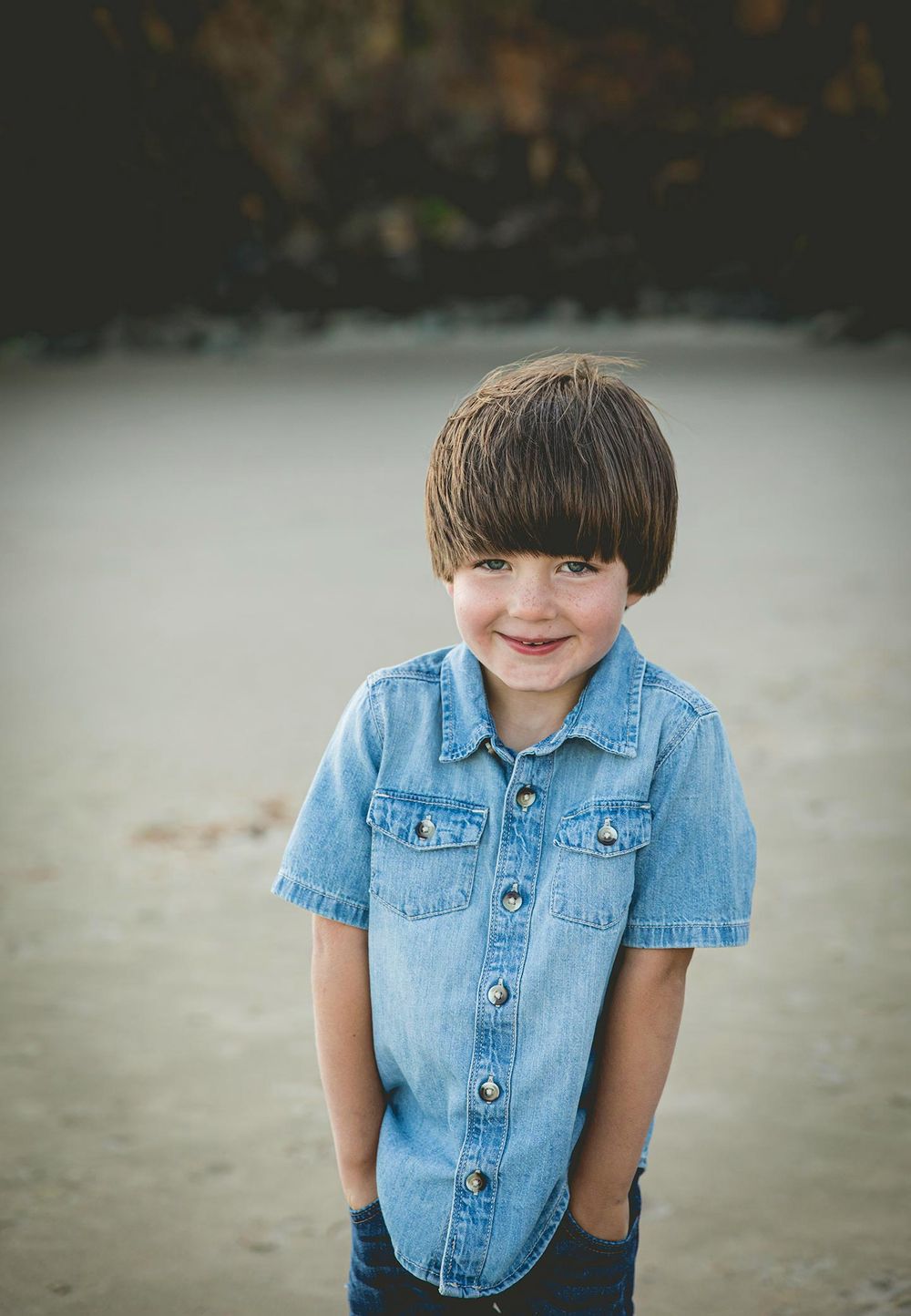 toddler boy wearing a denim button up shirt with his hands in his pockets on the beach