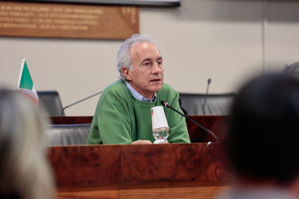Conference speaker in green sweater addressing attendees from wooden podium.