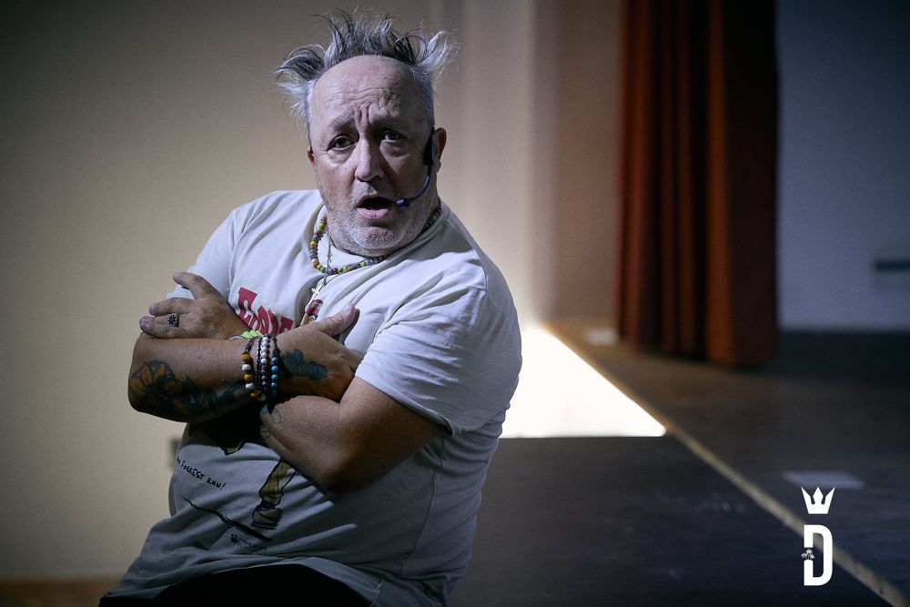 Theatrical performer in white t-shirt poses dramatically against dark background.