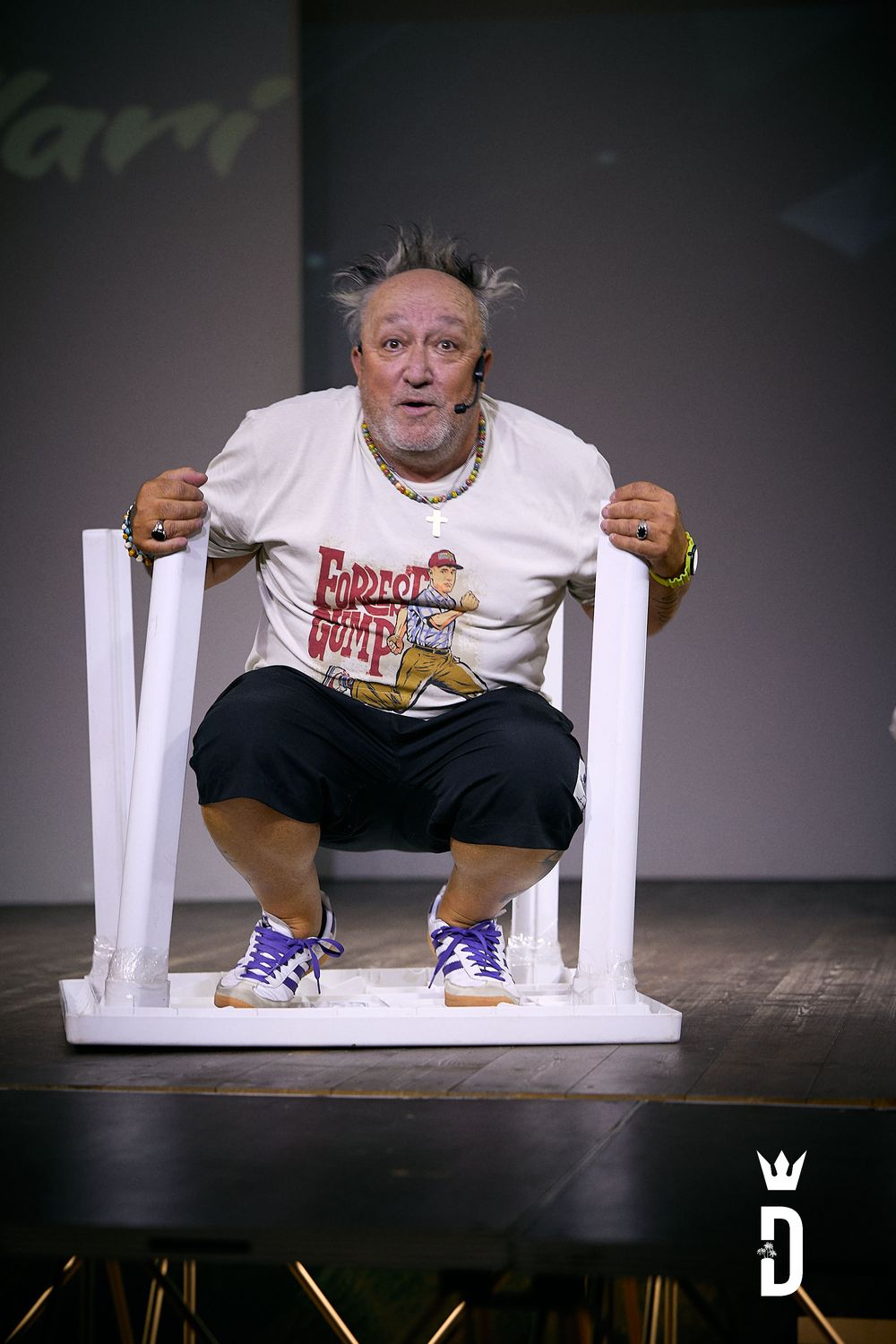 Performer squats on white chair during theatrical performance wearing printed t-shirt.