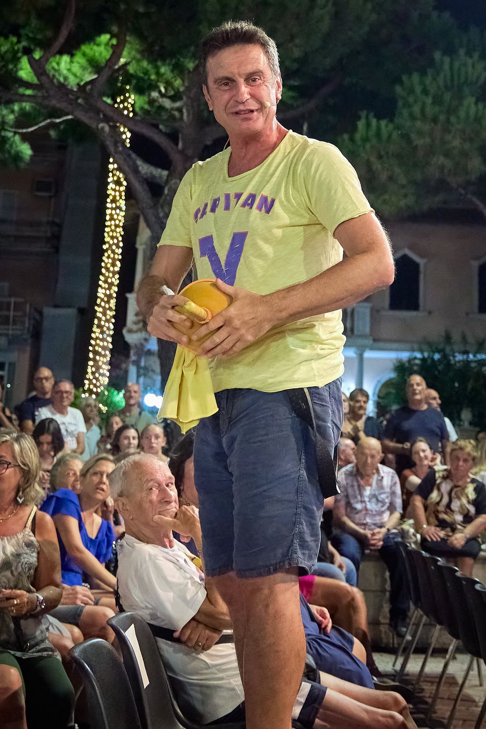 Stage performer in yellow t-shirt and shorts entertaining an outdoor audience.
