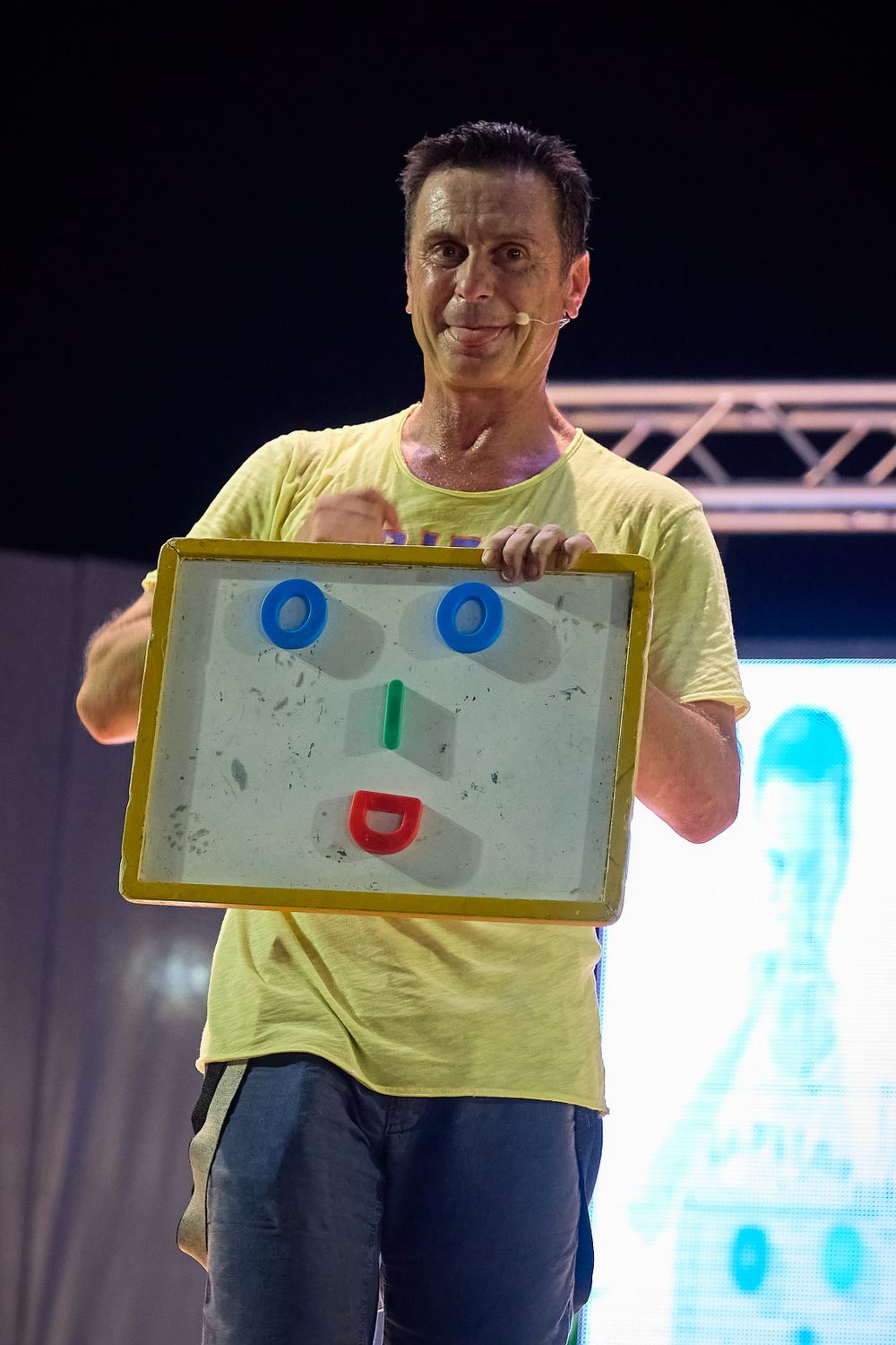 Performer in yellow shirt holding up a whiteboard with simple face drawing during stage show.