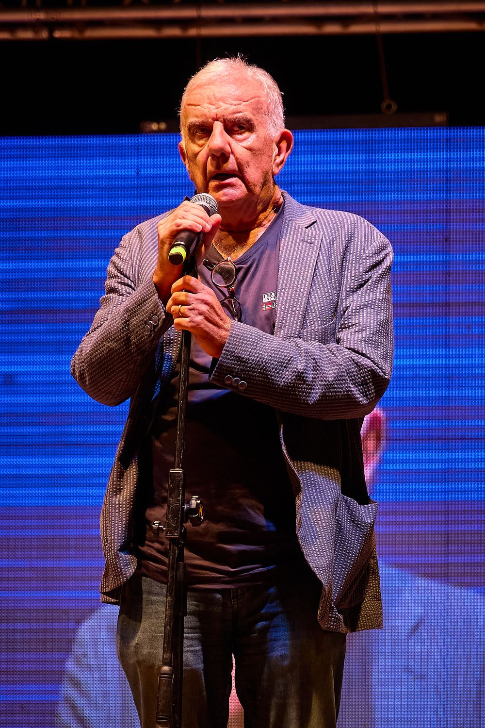 Speaker in blue blazer performing with microphone against illuminated blue background.