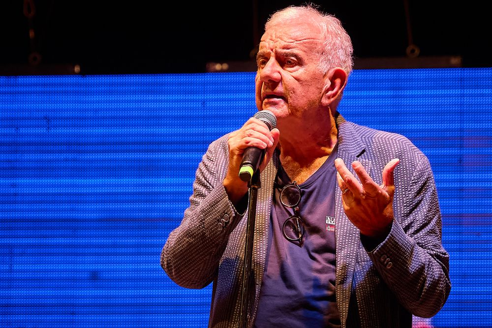 Performer speaking passionately into microphone against blue-lit backdrop on stage.