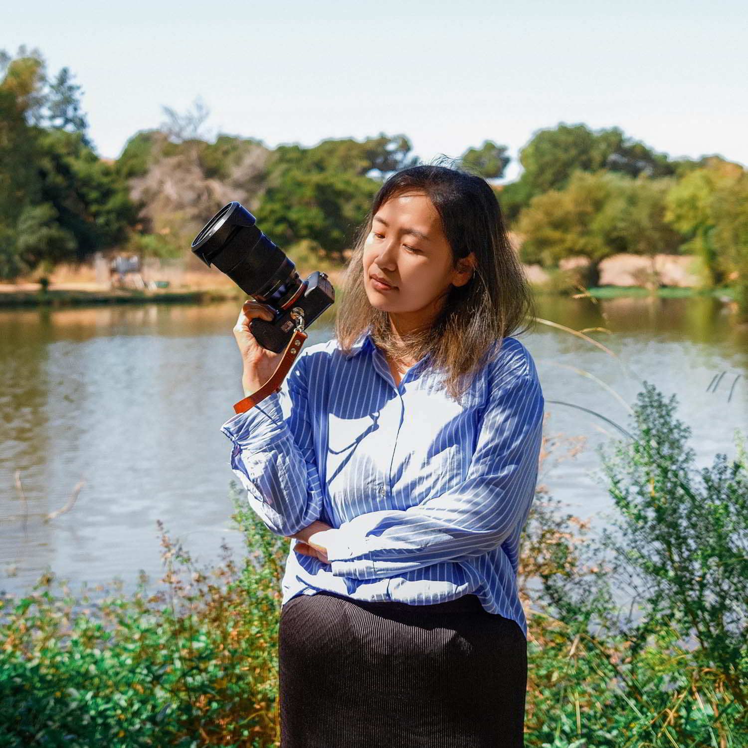 A photographer in a striped shirt stands by a scenic lake on a sunny day holding a digital camera.