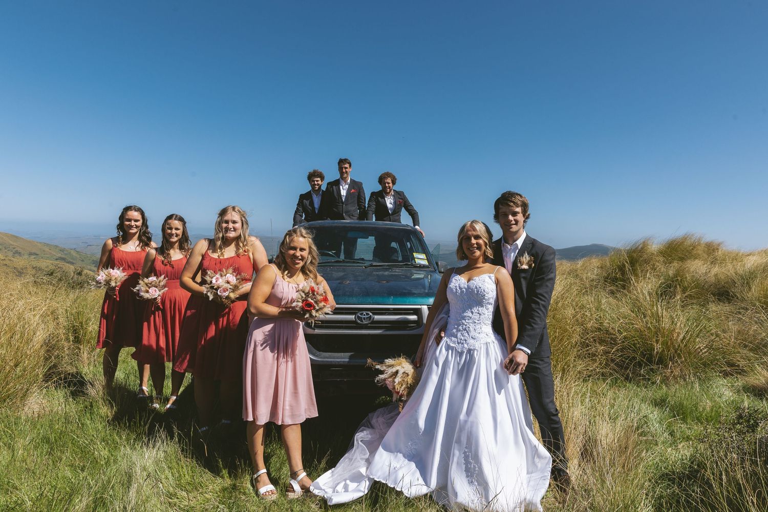 A wedding party poses with a vintage blue car on a scenic hilltop with green grass and blue skies.