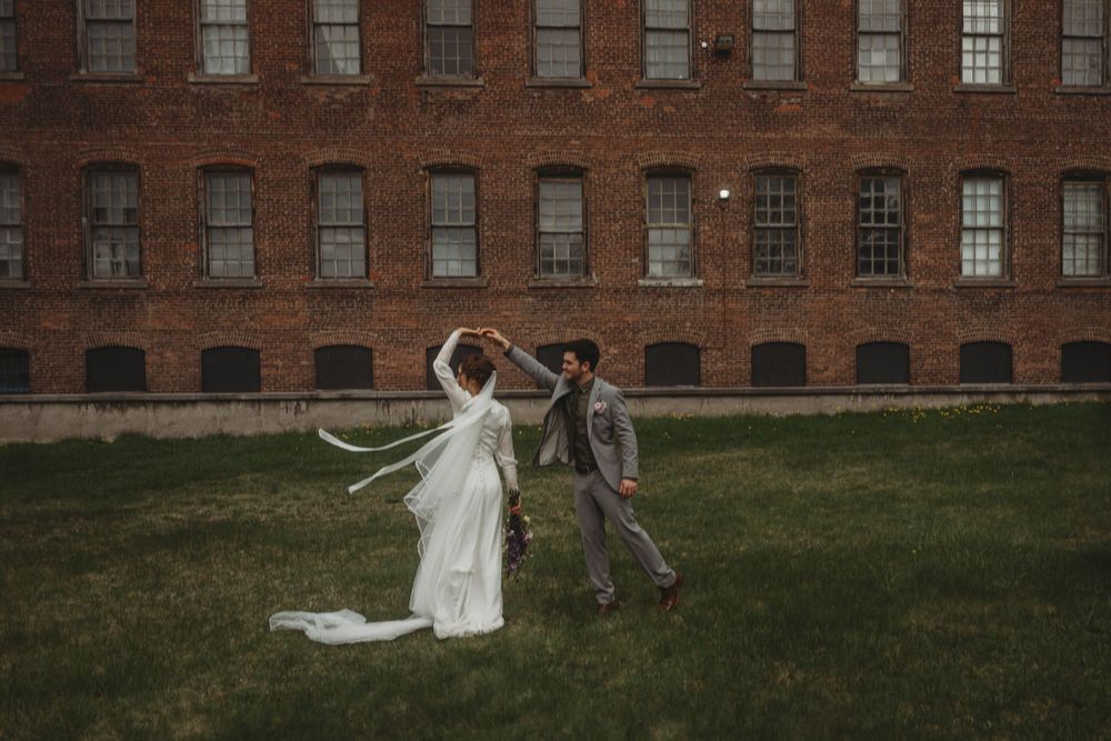 Wedding couple dancing playfully on lawn in front of historic brick building.