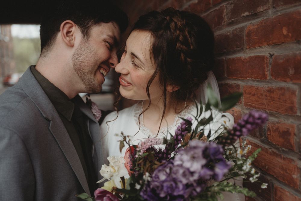 Romantic wedding couple sharing an intimate moment against rustic brick wall with purple floral bouquet.