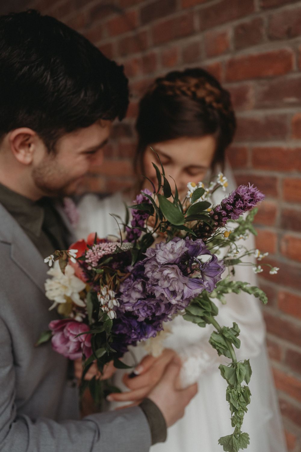 Close-up of wedding bouquet with purple and white flowers held between couple against brick wall.