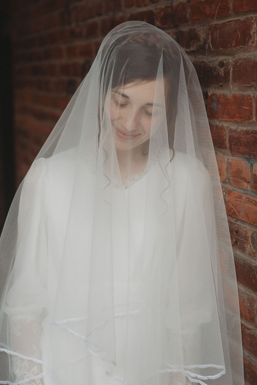 Ethereal bridal portrait with flowing white veil against rustic brick backdrop.