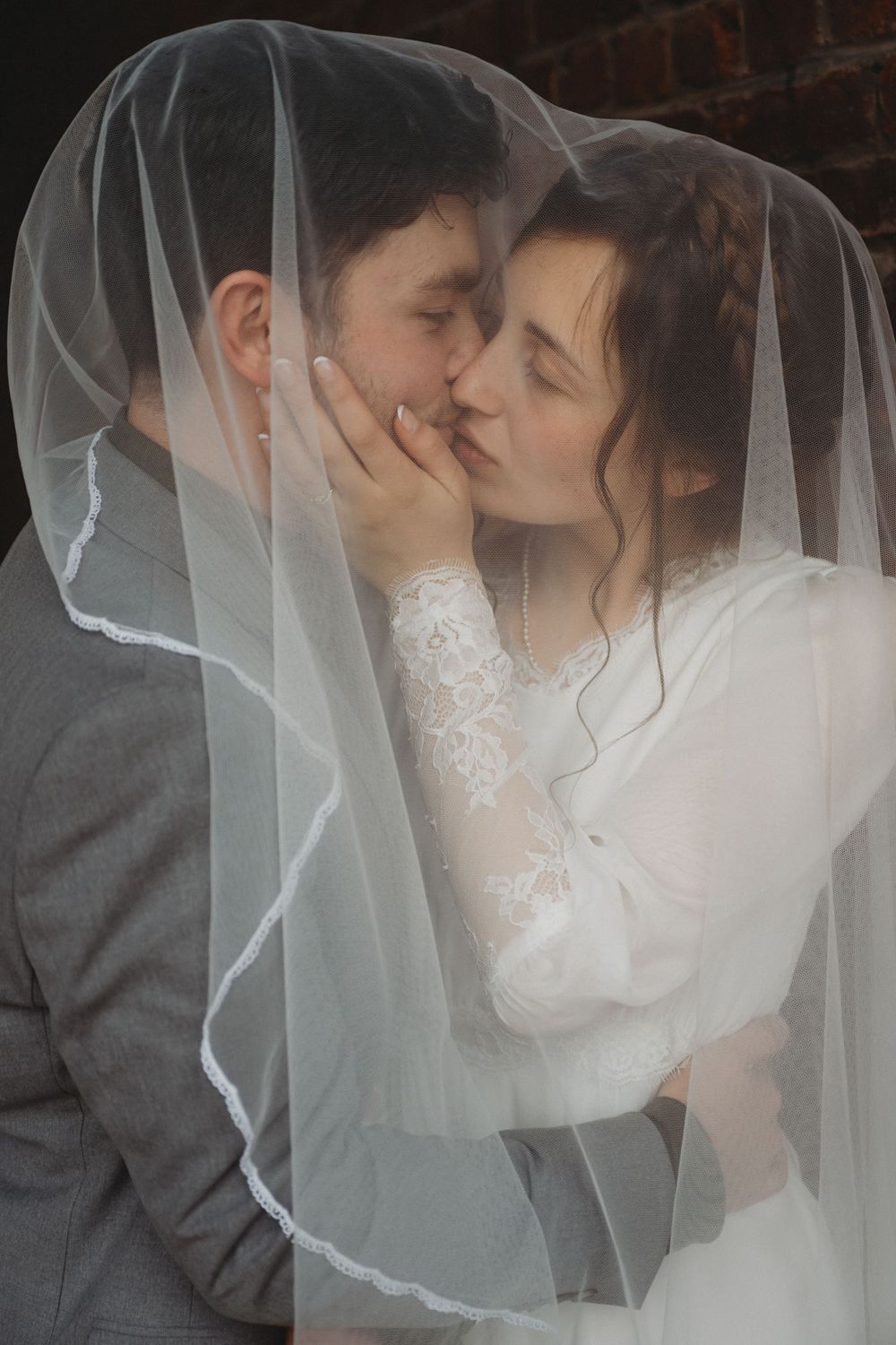 Romantic moment under flowing white veil against brick wall background.