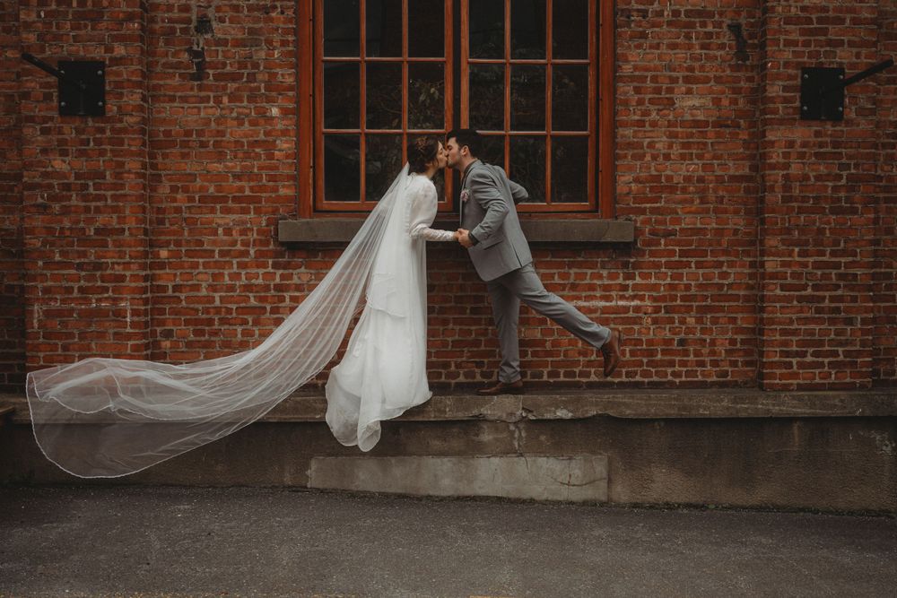 Creative wedding photo with long flowing veil against industrial brick building.