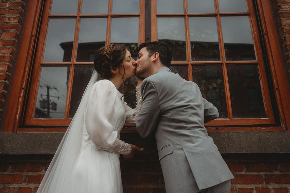 Intimate wedding kiss against vintage brick wall with orange window frame.