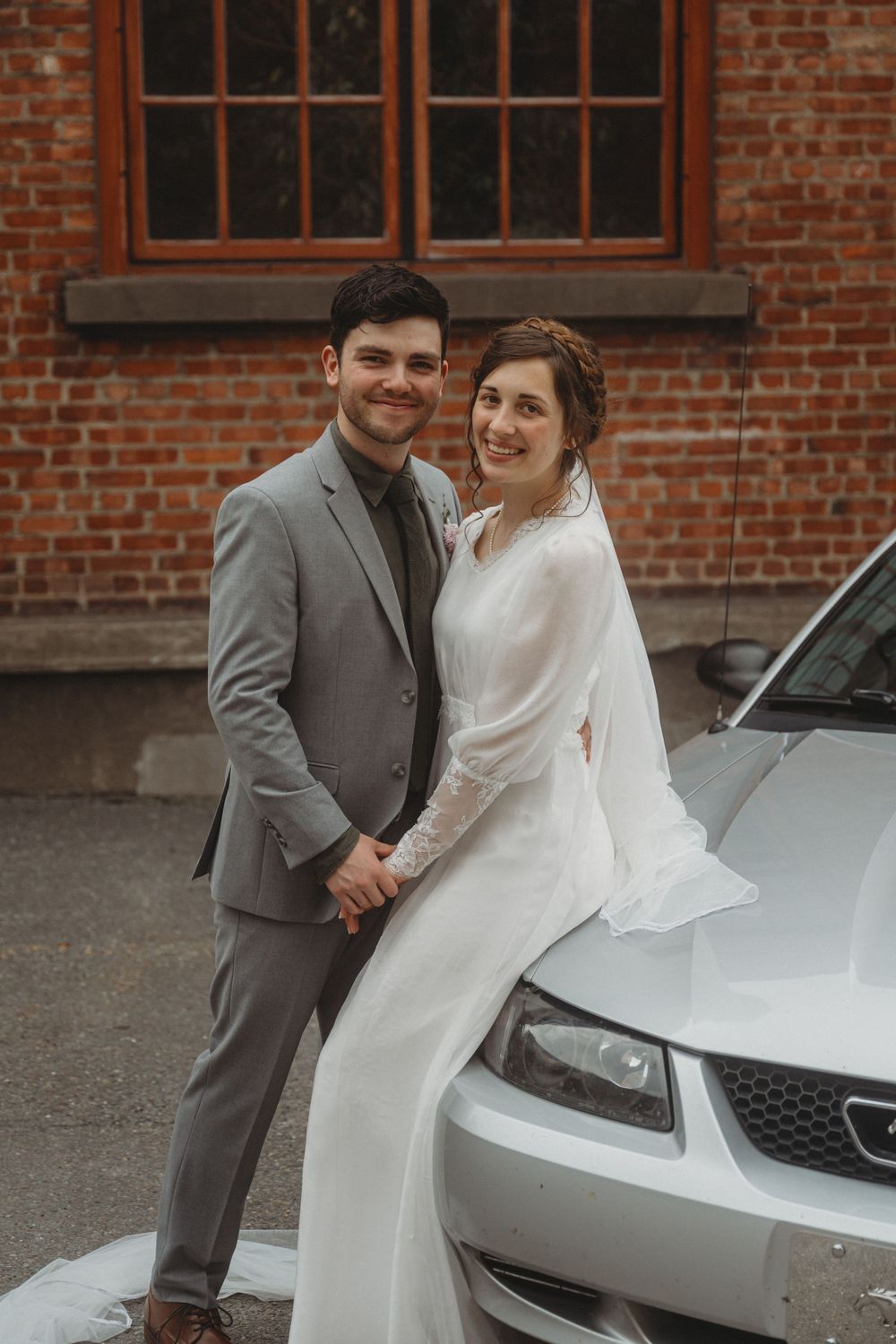 Newlyweds in grey and white attire pose beside silver car against brick building.