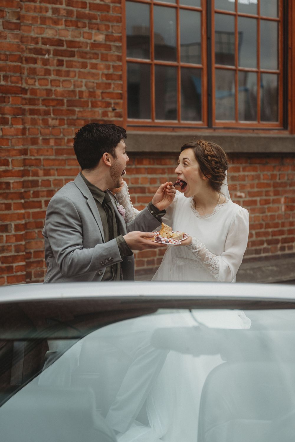 Wedding couple sharing cake in front of car against brick building backdrop.