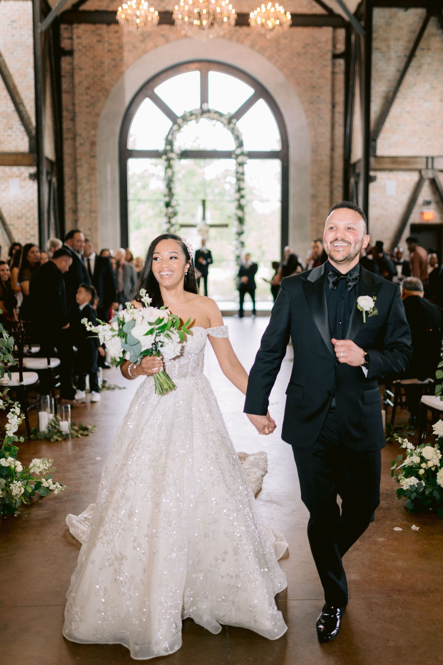 Newlyweds exit their ceremony under elegant chandeliers and arched window.