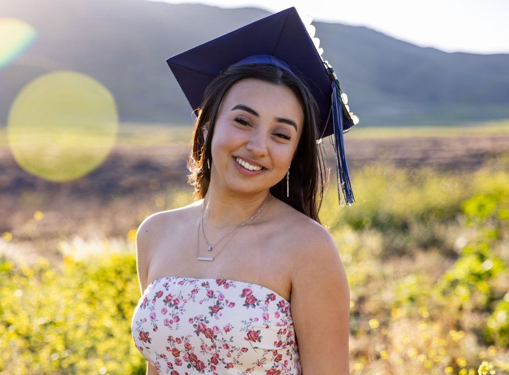 Graduate in cap and gown poses in floral dress against mountain backdrop at sunset.