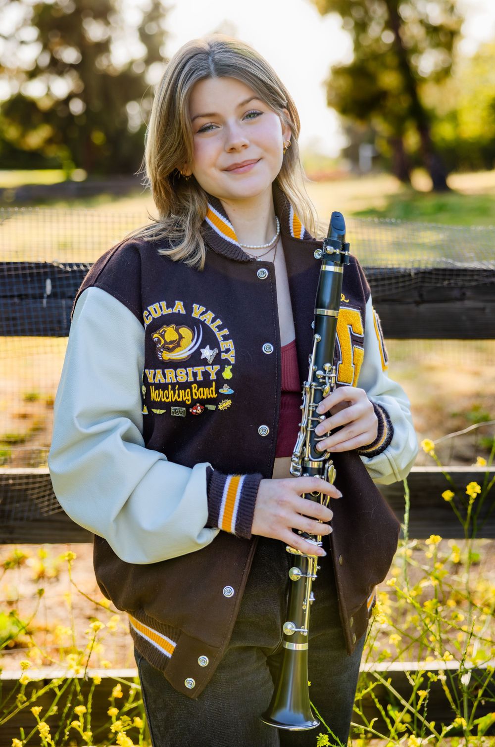 High school student in varsity letterman jacket holds a clarinet outdoors near wooden fence.