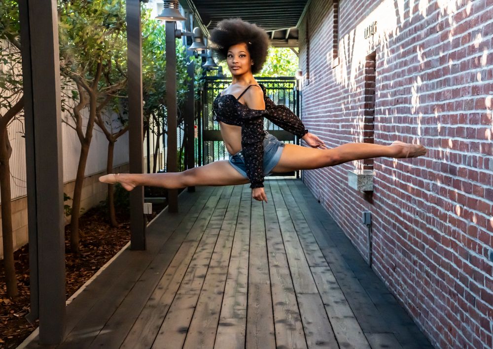 Dancer performing a split leap on wooden deck with brick wall backdrop.