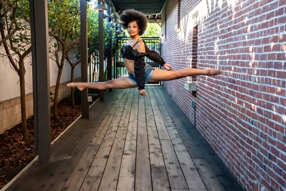 A dancer performs a graceful horizontal split pose between a brick wall and railing on a wooden deck.