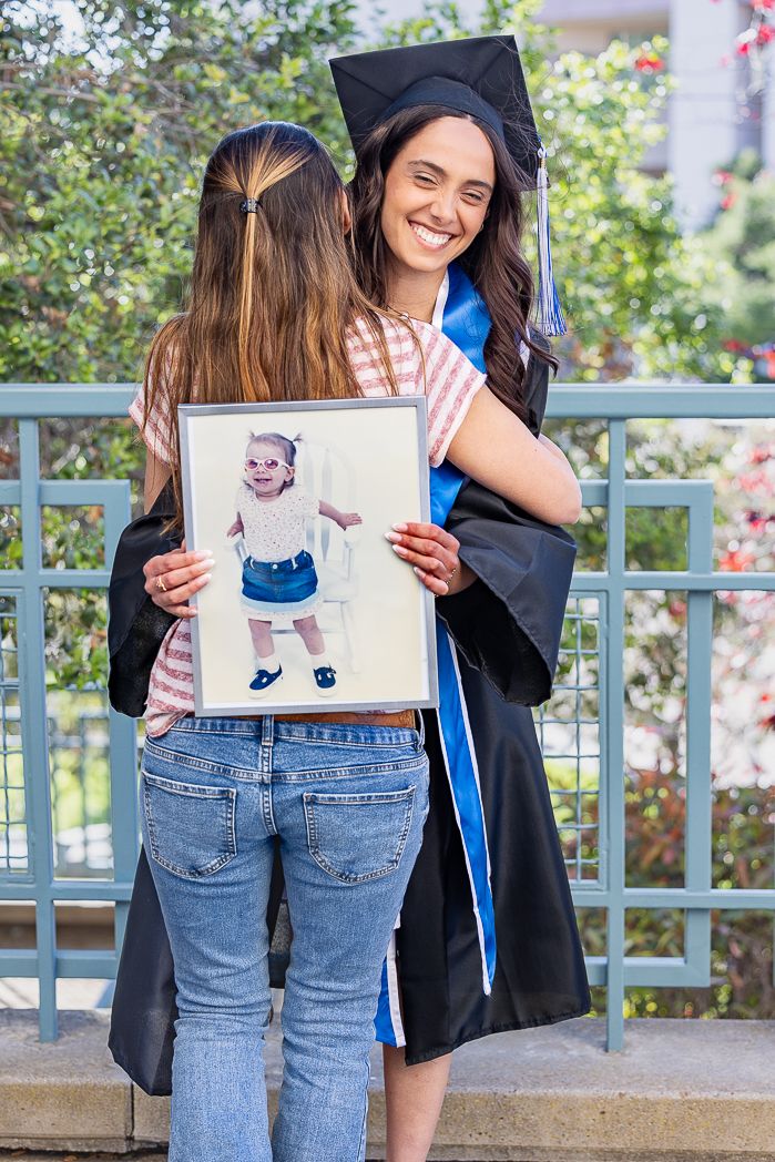 Graduate in cap and gown hugs someone holding framed photo during celebration.