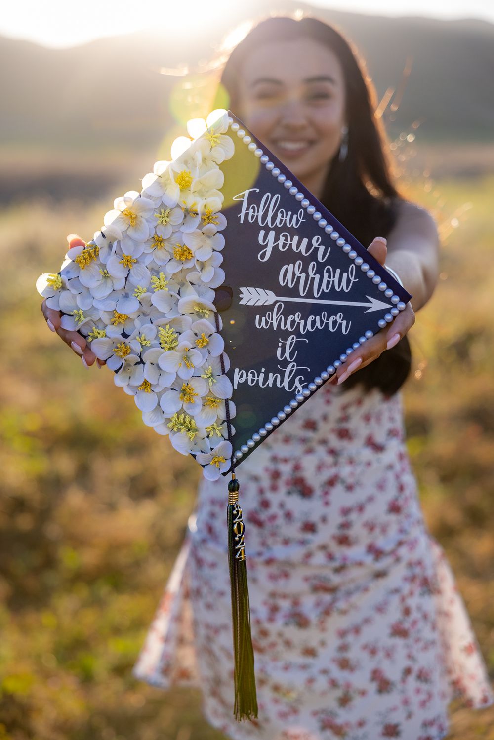 Decorated graduation cap with flowers and 'Follow Your Arrow' quote in sunlit field.