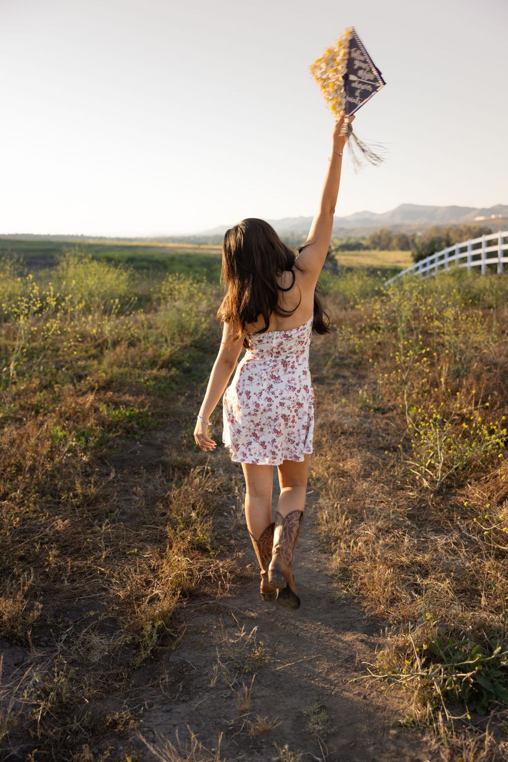 Graduate in floral dress holds decorated cap while walking on dirt path at sunset.