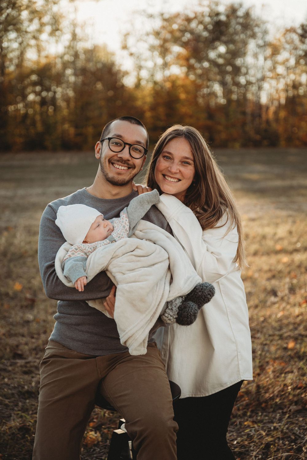 A couple holds their newborn baby while standing in a golden autumn field at sunset.