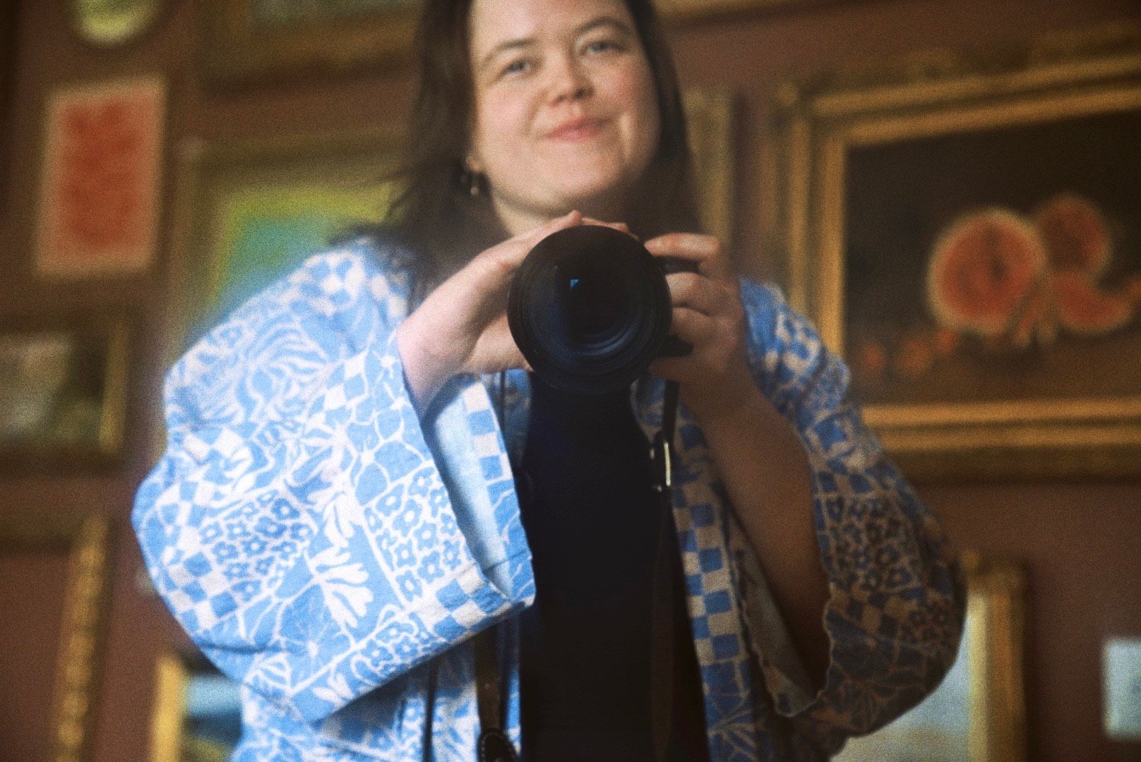 A photographer wearing a blue and white patterned blouse holds a professional camera in an indoor setting.