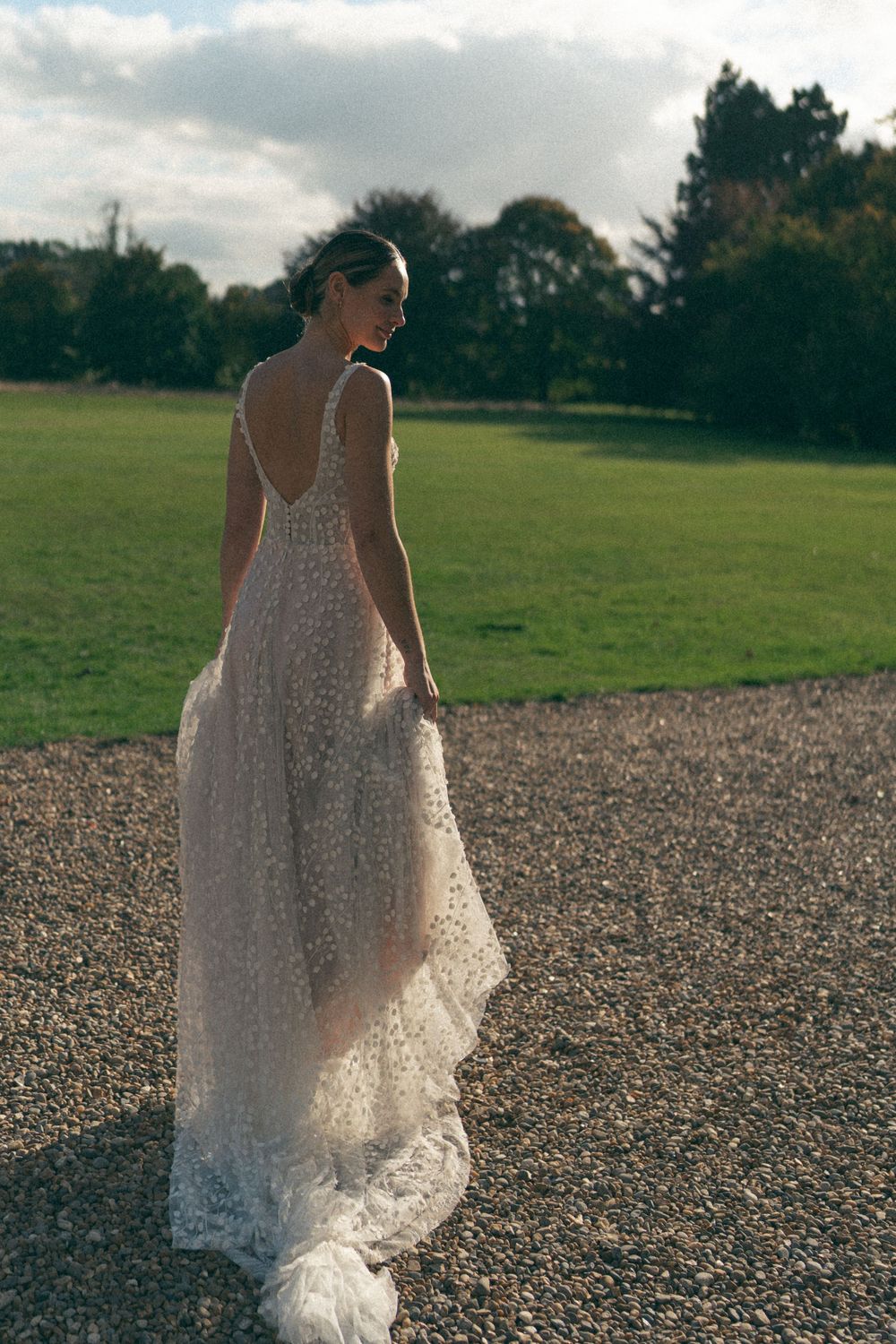 A person in a flowing white lace wedding dress stands on a gravel path next to a green field at sunset.