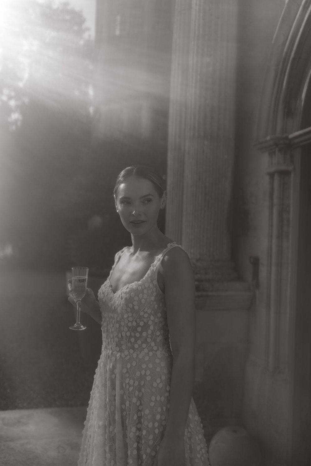 A dreamy black and white portrait of a person in a polka dot dress holding a wine glass by a column.