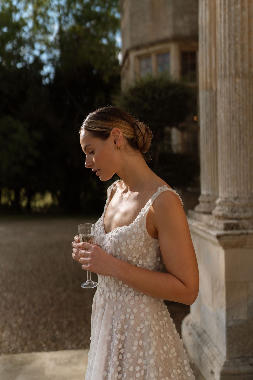A woman in a white lace wedding dress holds a champagne glass while standing by elegant stone columns in soft sunlight.