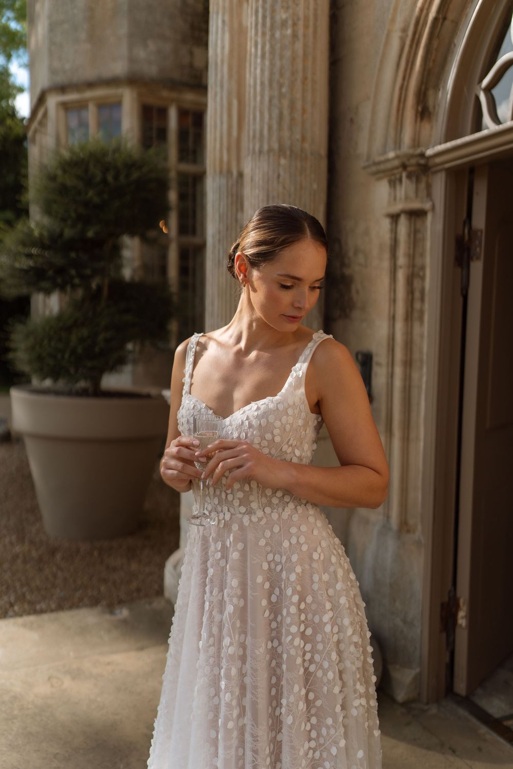 Woman in a romantic white lace wedding dress with spaghetti straps stands in front of stone architecture.