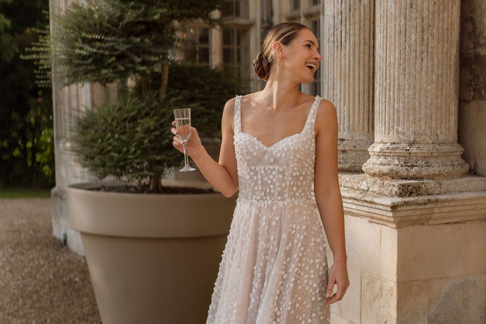 A white lace wedding dress is showcased against ornate stone pillars and greenery while holding a champagne glass.