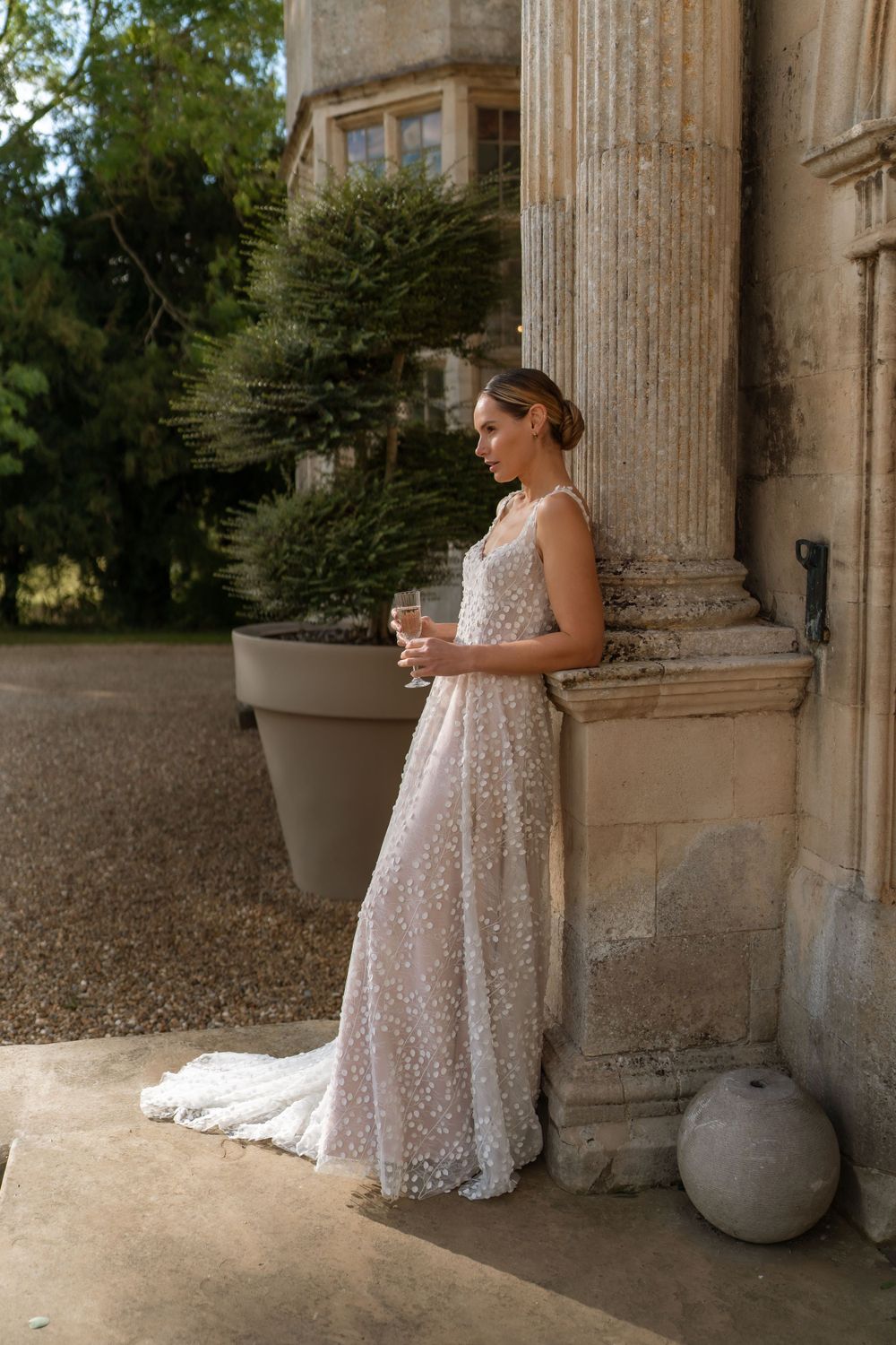 Elegant figure in a white lace wedding gown stands beside classic stone columns holding a coffee cup.