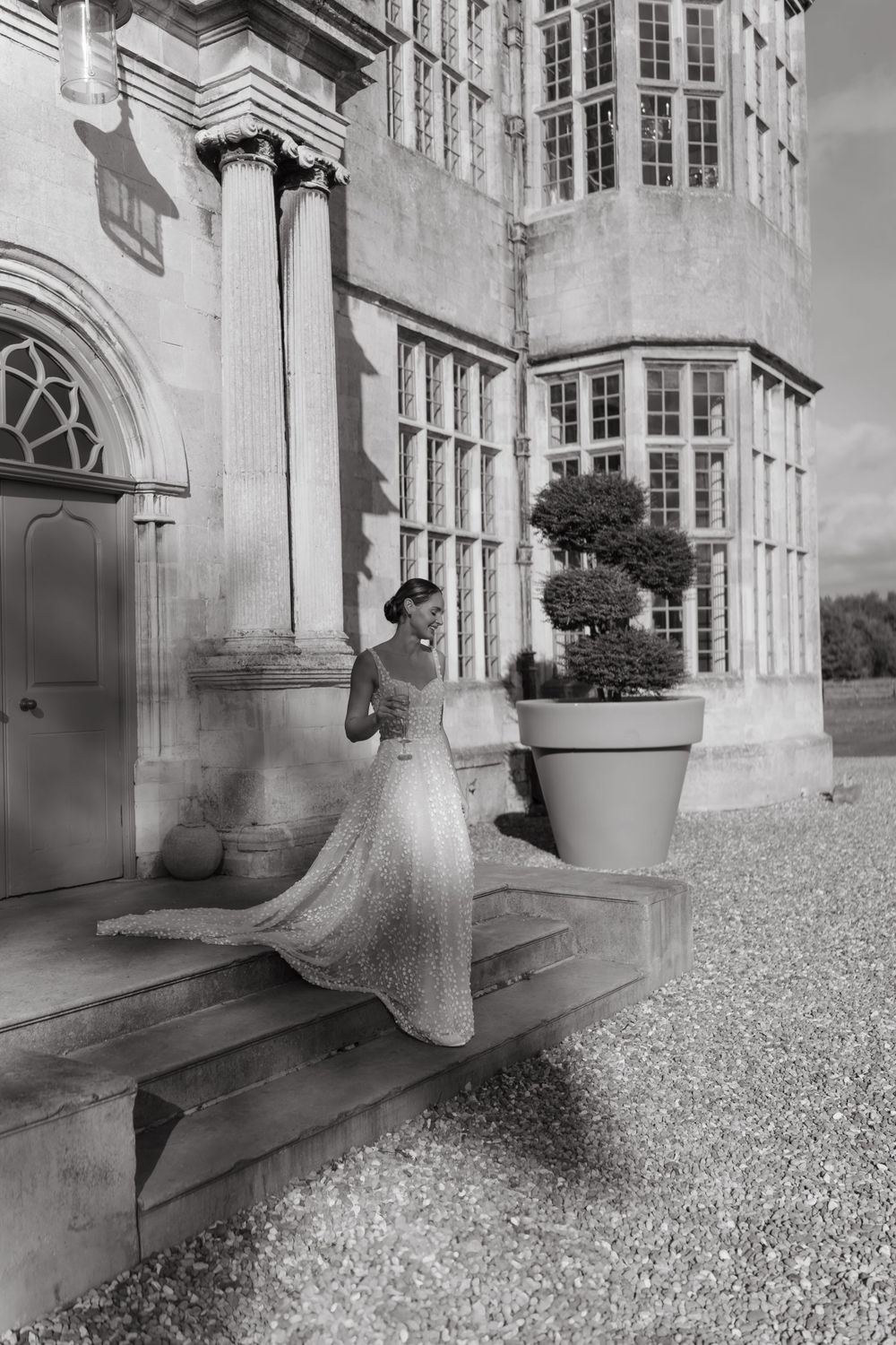 Black and white photo of a bride in flowing lace gown standing on stone steps of an elegant historic mansion.