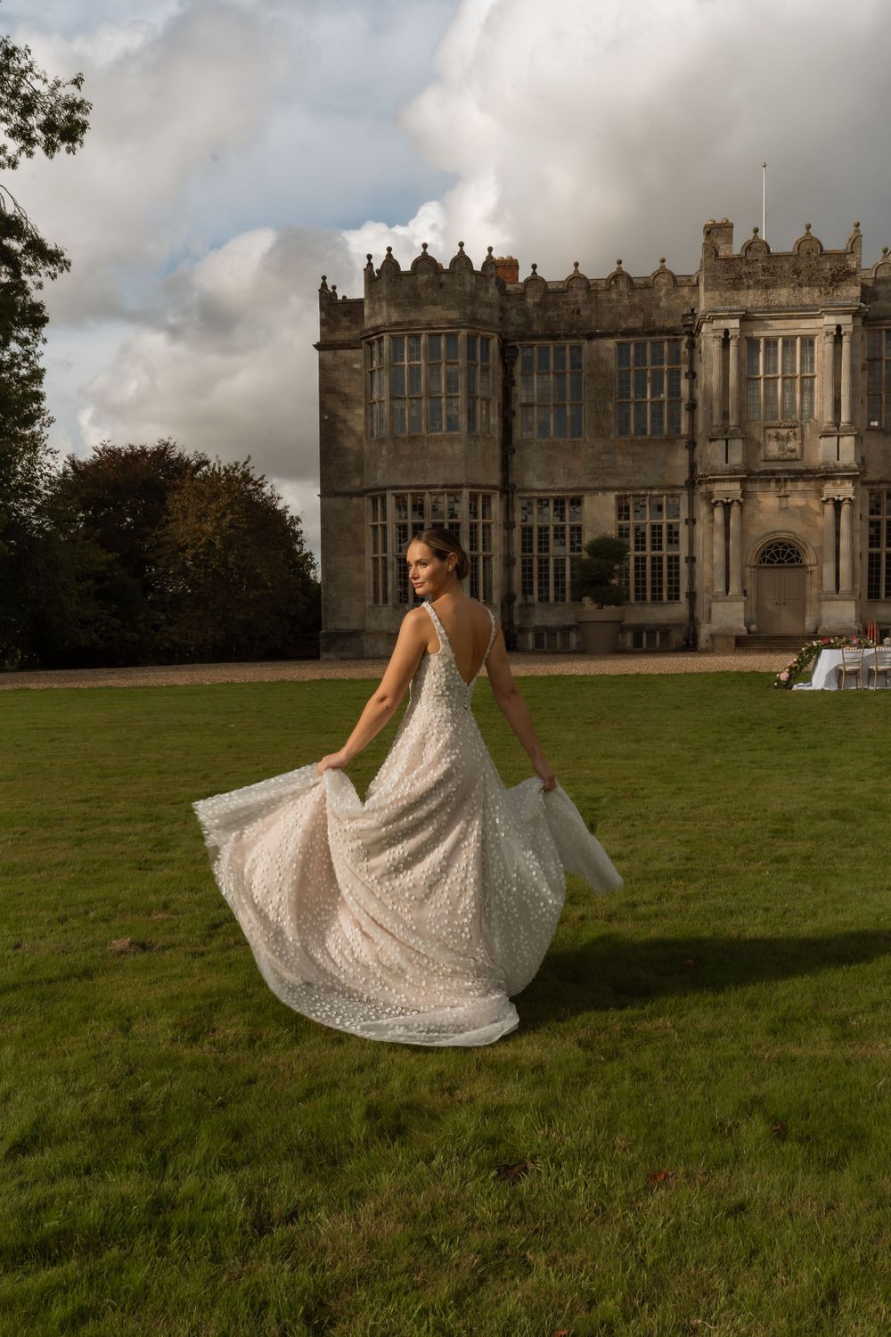 A sparkling white wedding dress twirls on a green lawn in front of a grand Gothic-style mansion with ornate windows.