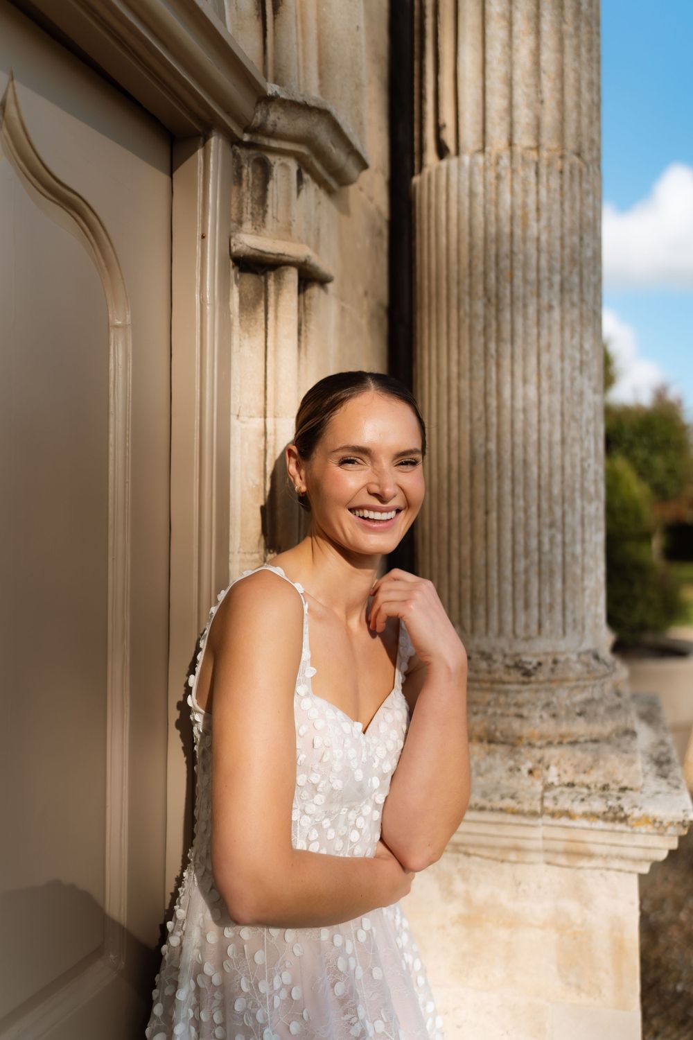A bride stands in a white lace gown by classical stone columns on a sunny day.