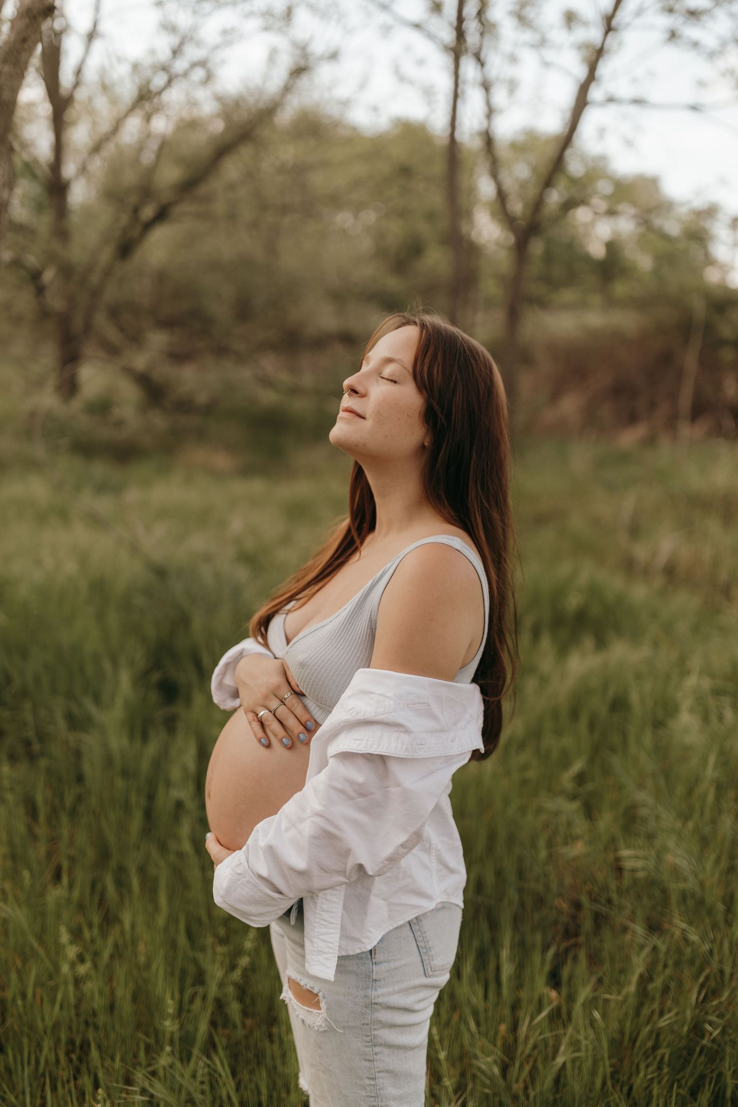 A pregnant woman in a white outfit stands serenely in a grassy field during golden hour.