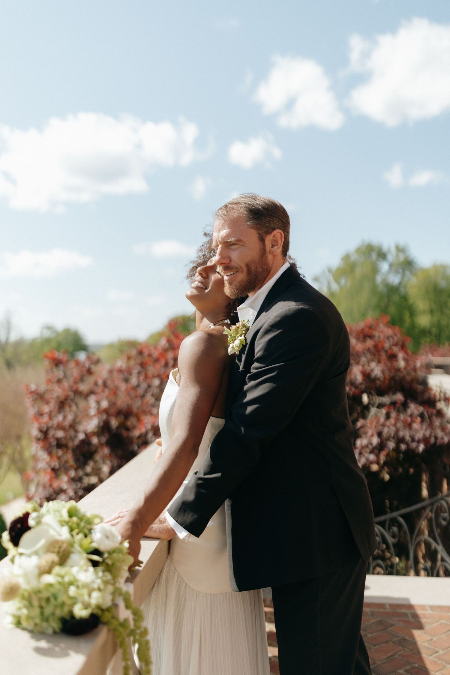 A couple in formal wedding attire embrace outdoors on a sunny day with flowering bushes and blue sky in the background.