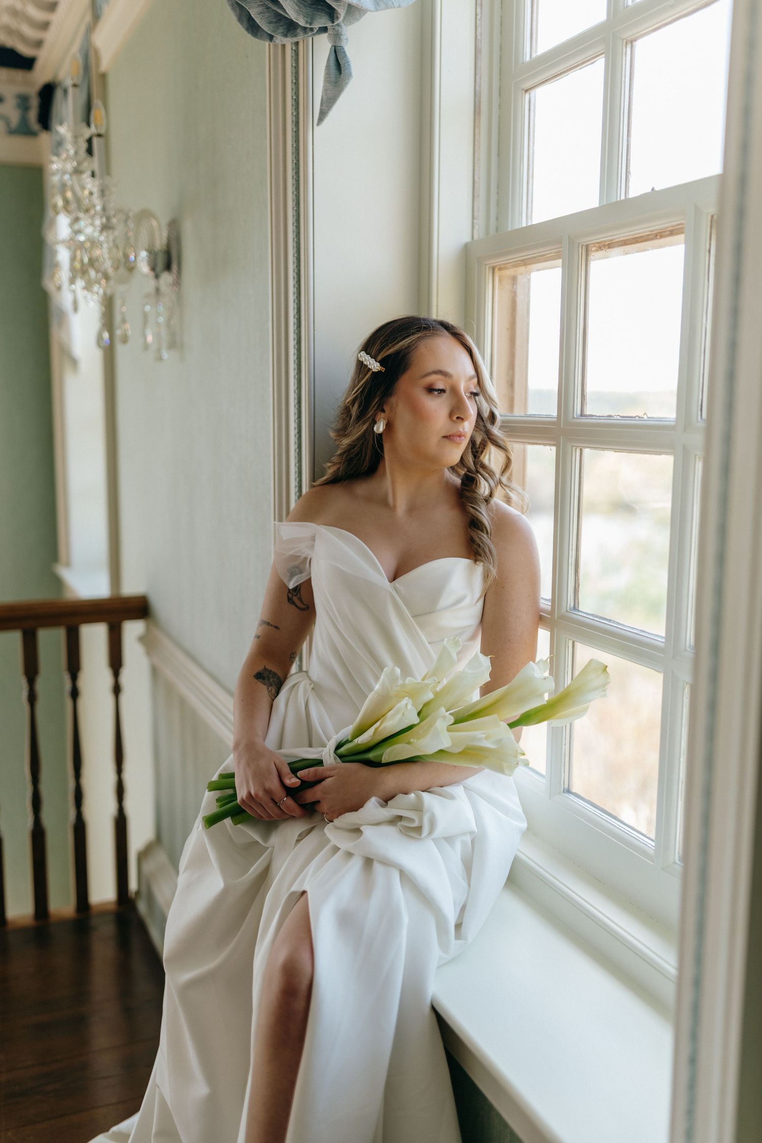 A bride in an elegant white off-shoulder gown gazes out a sunlit window while holding white calla lilies.