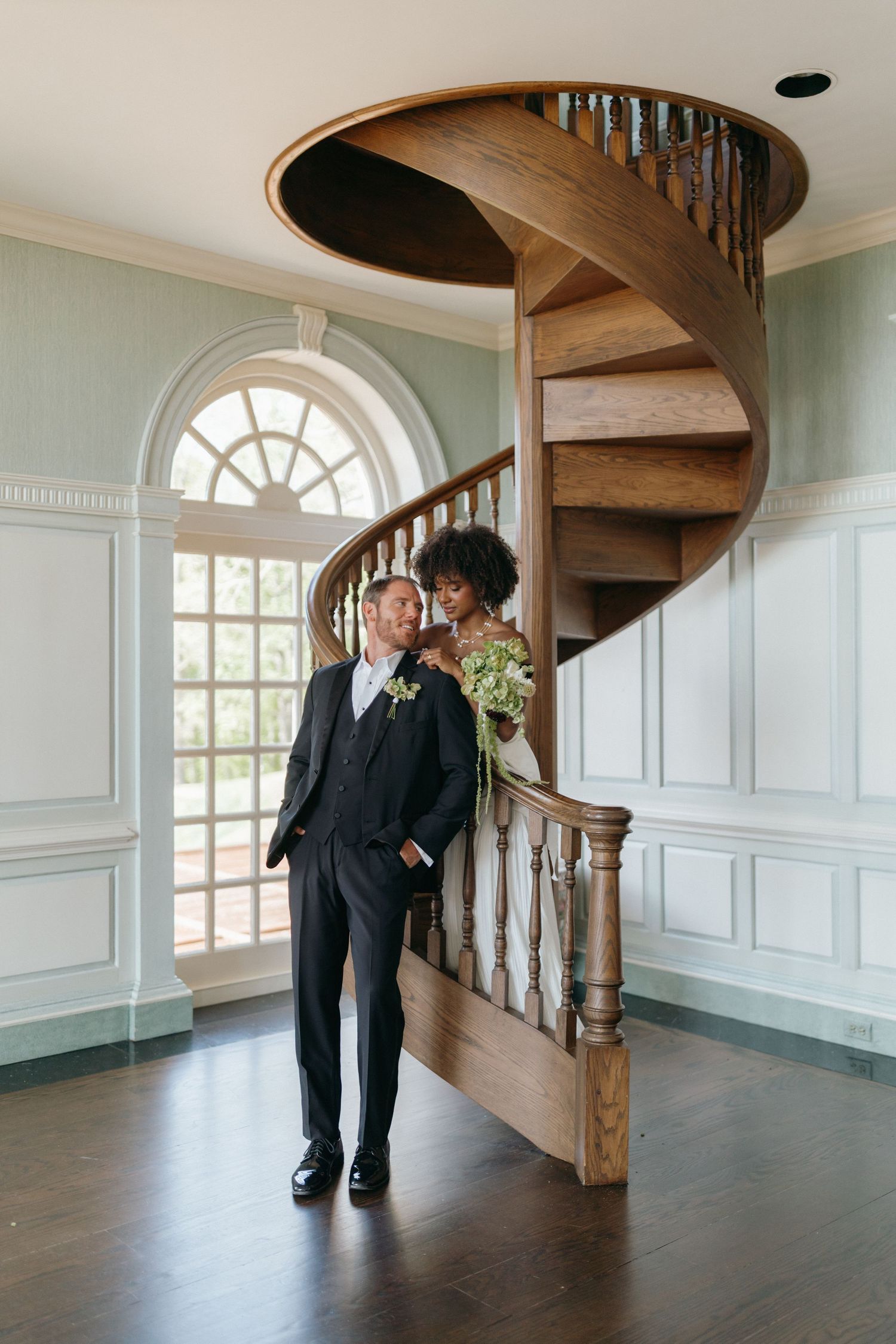 A couple embraces near an elegant wooden spiral staircase in a room with sage green walls and white wainscoting.