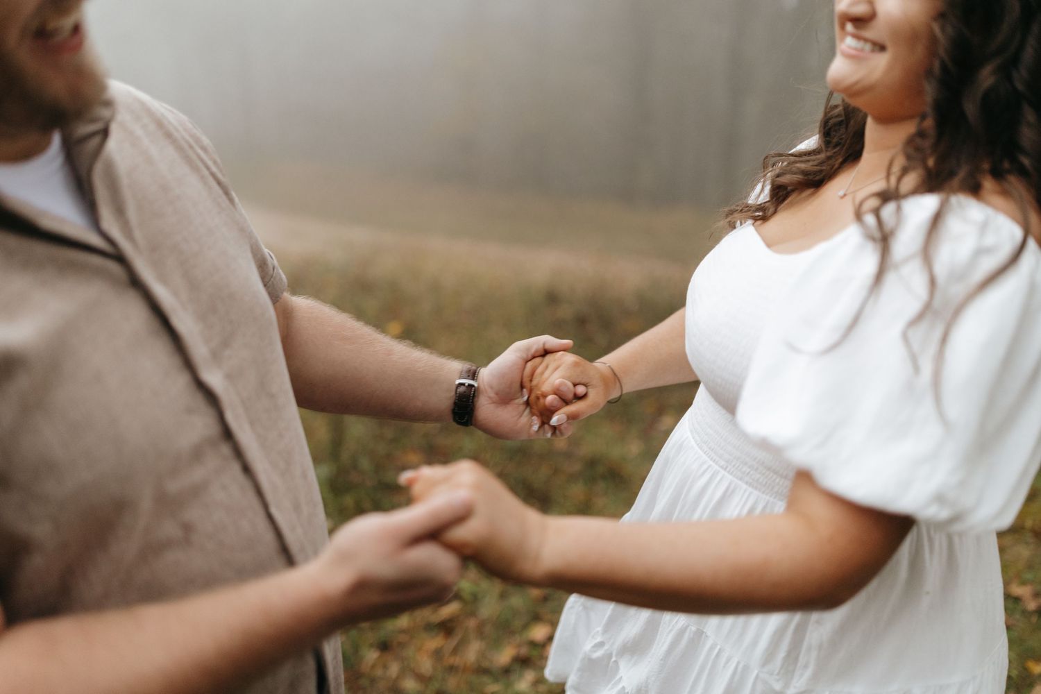 A couple holds hands intimately in an outdoor setting wearing neutral colors during a romantic moment.