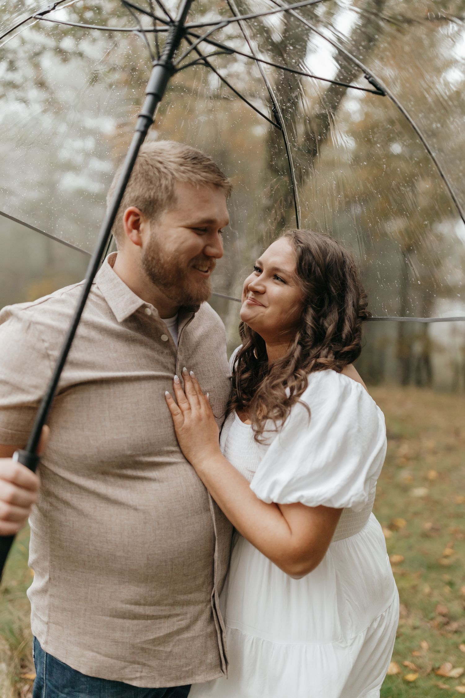 A couple shares a tender moment under a clear umbrella on an autumn day with fallen leaves in the background.