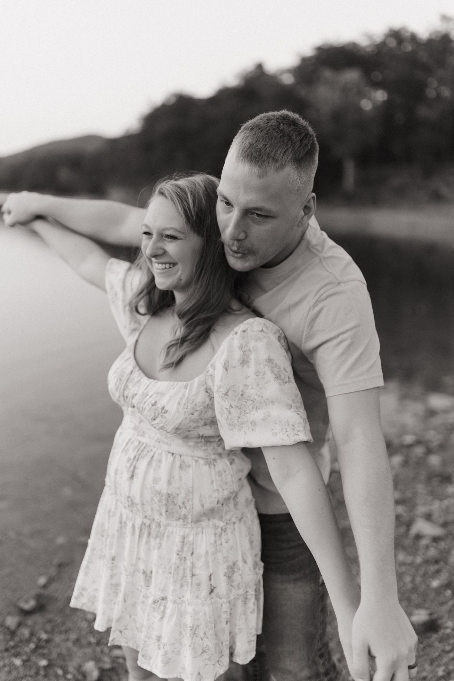 Couple embraces near water's edge in black and white photo with pregnant woman wearing floral sundress.
