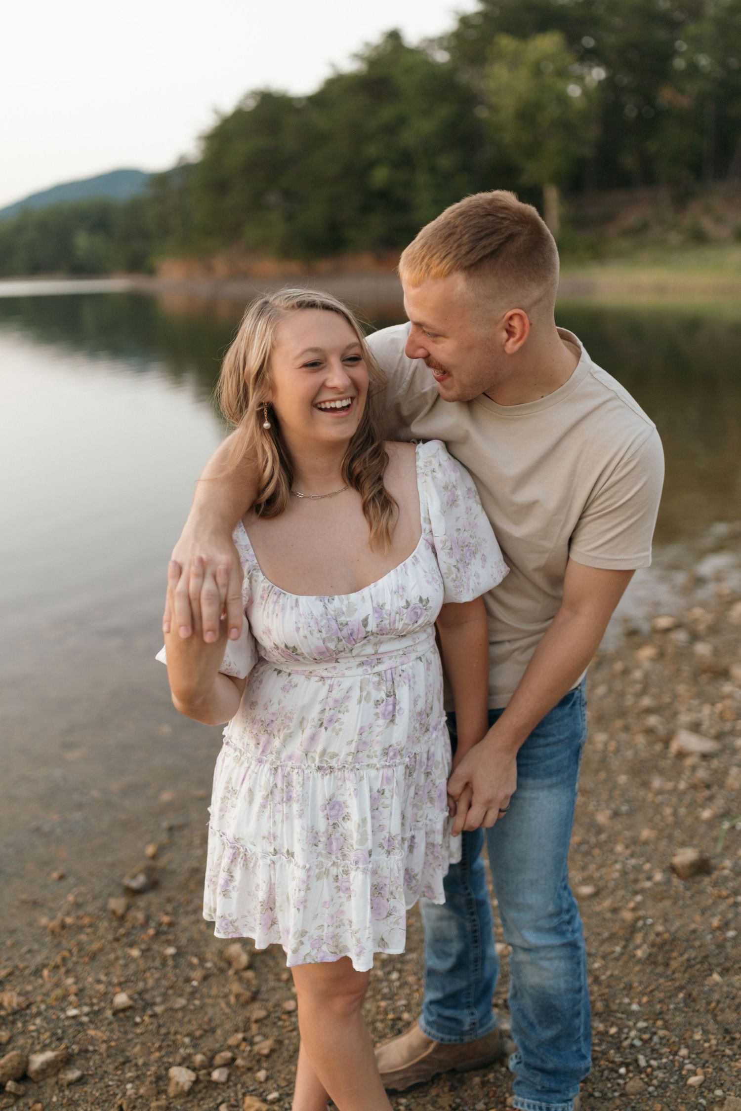 A couple shares a joyful moment together on a rocky lake shore at sunset.