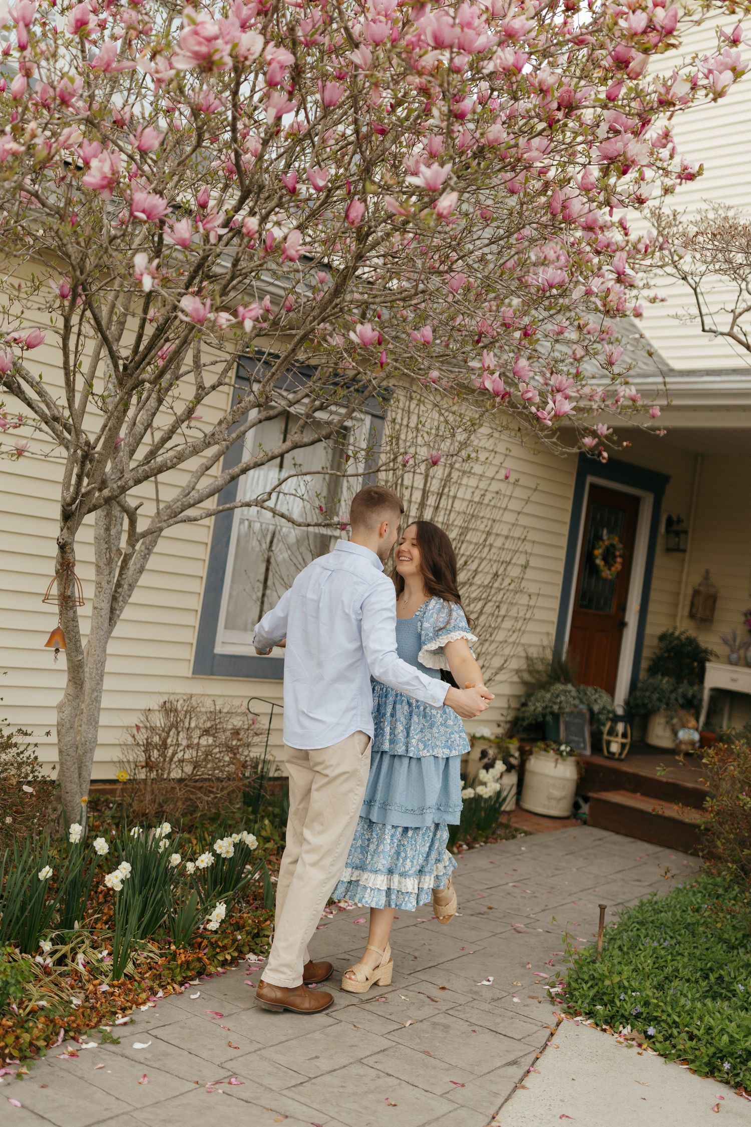 A couple shares a romantic moment under a blooming pink magnolia tree on a brick porch of a beige house.