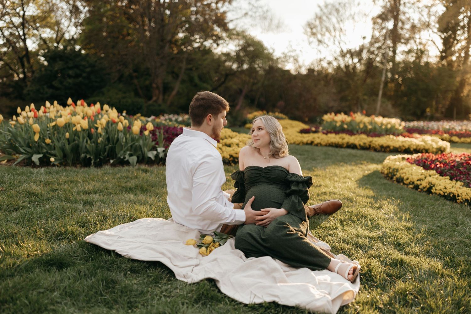 Couple having romantic sunset picnic in garden with colorful flowers and white blanket spread on grass.