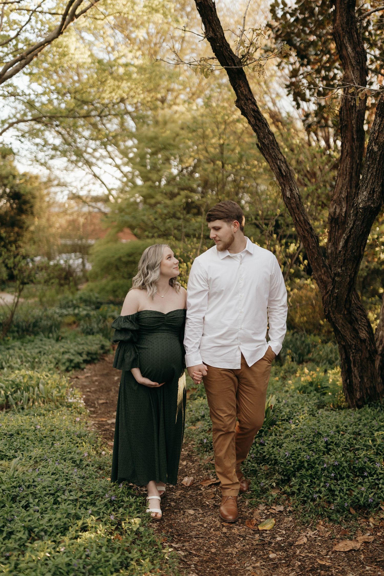 A couple walks together on a wooded path during autumn, with the expecting mother wearing an elegant dark green dress.