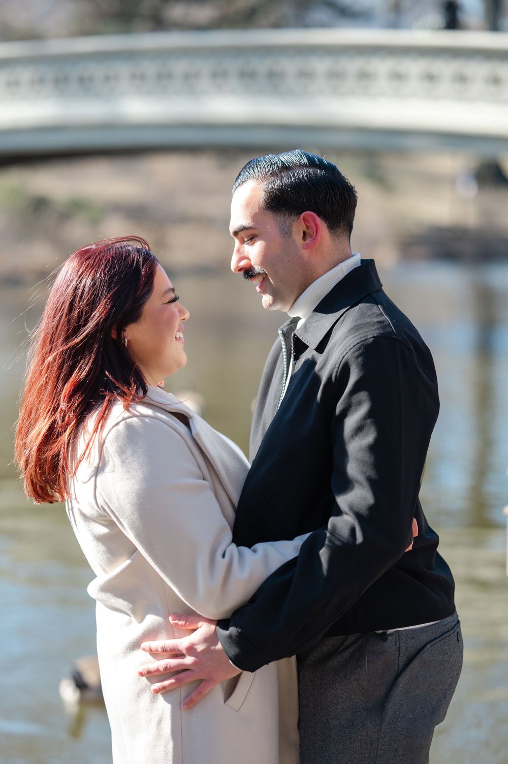 A couple shares a romantic moment by the water, with the woman in white and man in black formal wear embracing in front of Bow Bridge